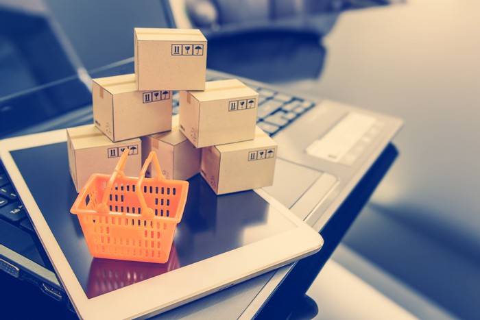 A miniature shopping cart and mini boxes laid atop a smartphone and laptop. 